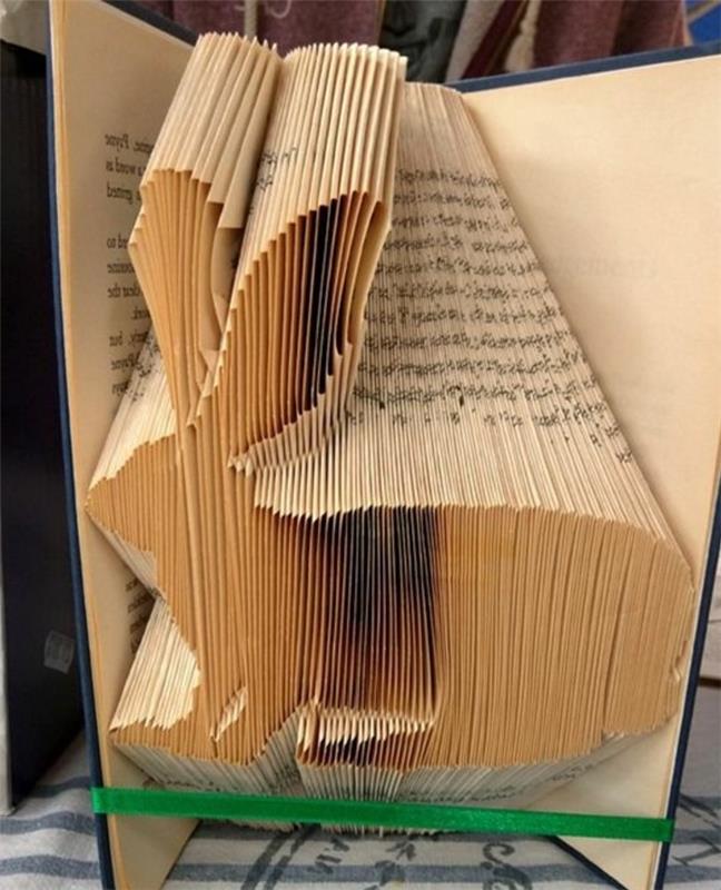 zloženi knjižni umetniški vzorci, v obliki zajca iz prepognjenih rumenih strani, znotraj odprte knjige s temno modrimi trdimi platnicami, vezane z zelenim trakom