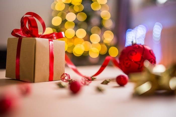 Kalėdinis fonas kaip dovana iš kraftpopieriaus, papuoštas raudona juostele, raudonu Kalėdų kamuoliu ir Kalėdų eglute, apšviesta šviesiomis girliandomis