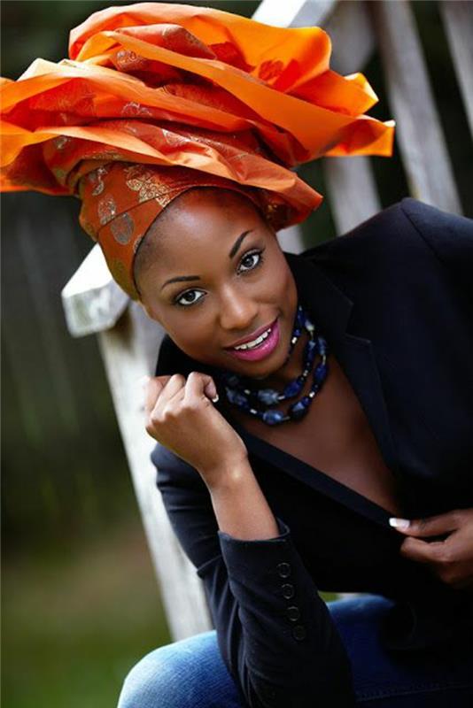 oranžinis turbanas, susietas su gėlėmis su dideliais žiedlapiais, afrikietiška apranga, afrikietiškas raštas, švarko juoda gili iškirptė, karoliai su ametisto akmenimis