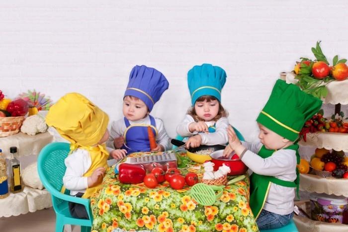 štirje malčki, ki nosijo kuharske klobuke in predpasnike v različnih barvah, sedijo na majhnih plastičnih stolih okoli mize z različnimi kuhinjskimi pripomočki in zelenjavo