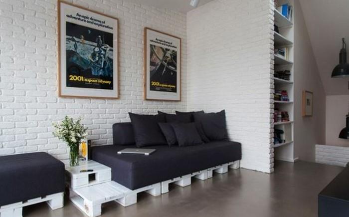 plytų siena balta, svetainės viduje su padėklų sofa, pagaminta iš baltų medinių padėklų, padengta juodomis sofų pagalvėlėmis