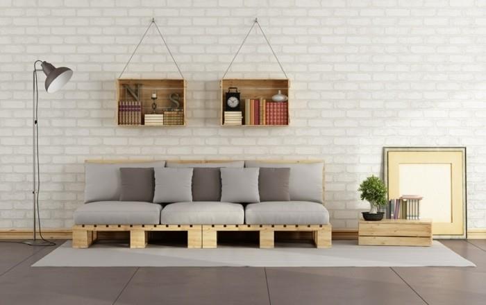 dvi medinės knygų lentynos, pakabintos ant baltos plytų sienos, virš sofos iš šviesių medinių padėklų ir padengtos pagalvėlėmis, šviesiai ir tamsiai pilkos