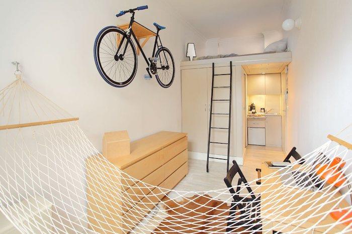 baltos sienos ir minimalistinis dizainas 20 m2 studijoje, studijos virtuvėlė ir drabužių spinta su lova aukščiau, vidinis hamakas, stalas ir medinė komoda