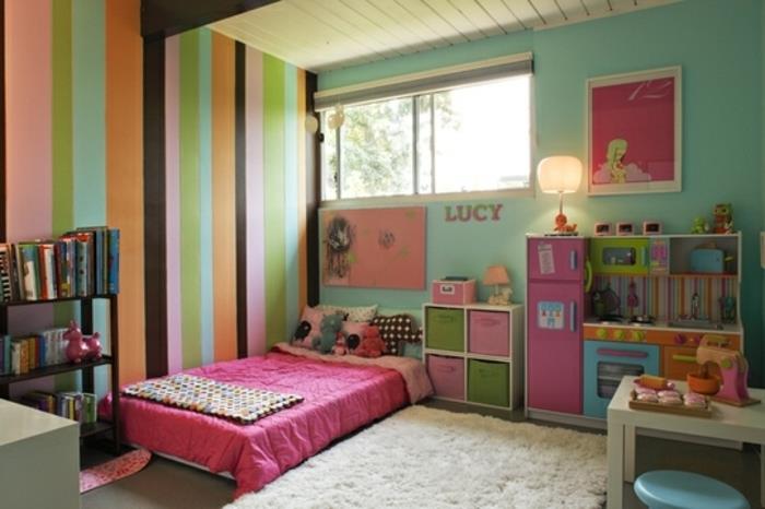 žalia siena ir akcentinė siena su įvairiaspalvėmis juostelėmis, lova ant grindų, rožinis lovos užtiesalas, beveik baltas kilimas, knygų spinta, maža virtuvė, saugykla, Montessori mergaitės kambarys