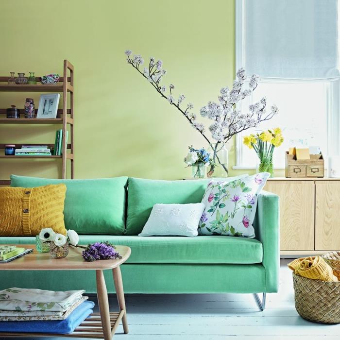jūra žalia sofa pistacijų žaliais dažais dekoruotoje svetainėje, balintos parketo grindys, medinis kavos staliukas, krepšelis ir medinė lentyna