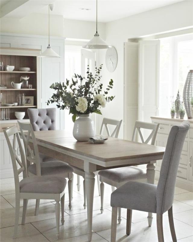 eleganten podeželski dekor v jedilnici, lesena miza, beli leseni stoli s sivo prevleko, vaza s cvetjem, bela komoda, viseče luči