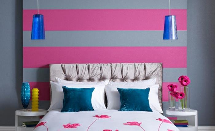 roza in siva spalnica, siva barva sten z rožnatimi črtami, sivo vzglavje, bela posteljna garnitura z rožnatimi cvetovi, modre blazine in vzmetenja