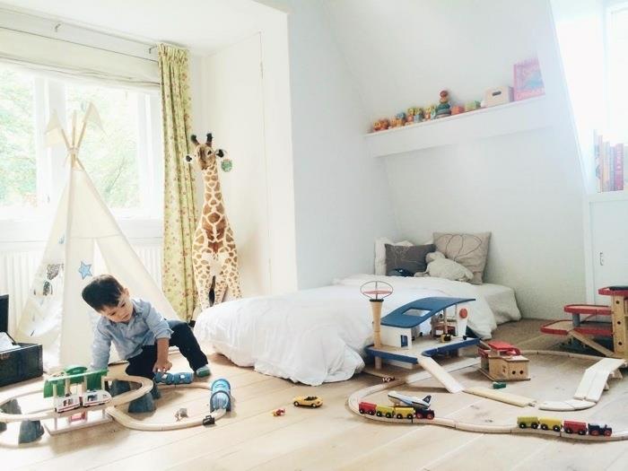 „Montessori“ pedagogikos vaikų miegamojo išdėstymas, šviesus parketas, vaikiškas marškinėliai, berniuko miegamojo dekoras, čiužinio kilimėlis ant grindų, žirafos pliušas, žaislų laikymo lentyna