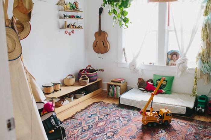 idėja, kaip sutvarkyti „Montessori“ miegamąjį, rytietišką kilimą, čiužinį ant grindų, baltą sieną, gitarą, žemą medinę saugyklą