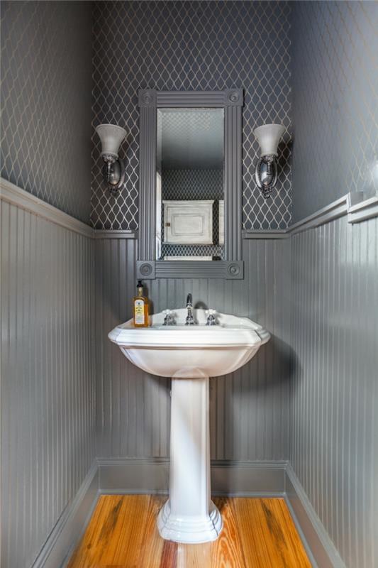 tualetas pilkos spalvos, baltas pjedestalo baseinas, du žvakidės ir įrėmintas veidrodis, geometriniai sienų tapetai, medinės grindys