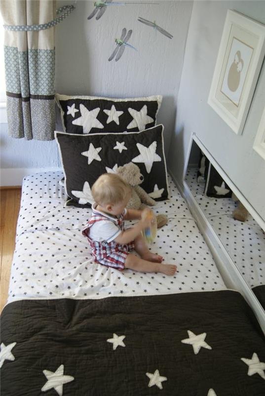 lovytės čiužinys ant grindų, baltas su juodais taškeliais, lovatiesė, juodos pagalvės su baltomis žvaigždutėmis, balta siena, kūdikis, meškiukas