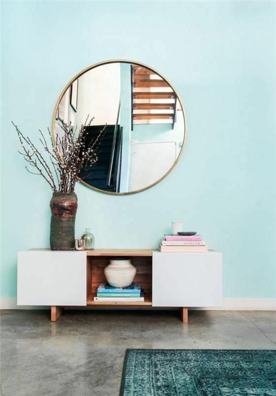 vandens žali dažai ant sienos, didelis apvalus veidrodis, laikymo spintelė, betoninė danga, anties mėlynas kilimas ir šakos vazoje