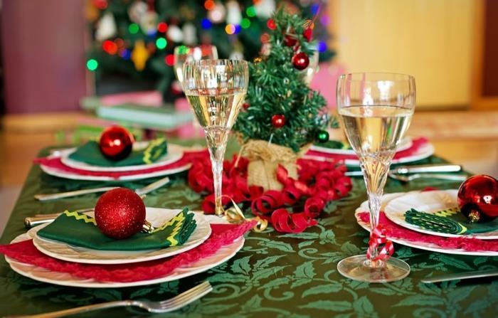 žalia staltiesė su gėlių raštais, mažas dekoratyvinis medis, raudoni dekoratyviniai rutuliai, žalios servetėlės, šampano taurės