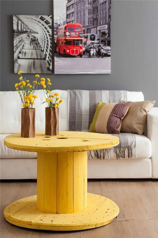 lesena bobnasta miza prebarvana v rumeno, izrezljane lesene vaze za rože, bela zofa in večbarvne blazine, okrasne plošče Londonske pokrajine