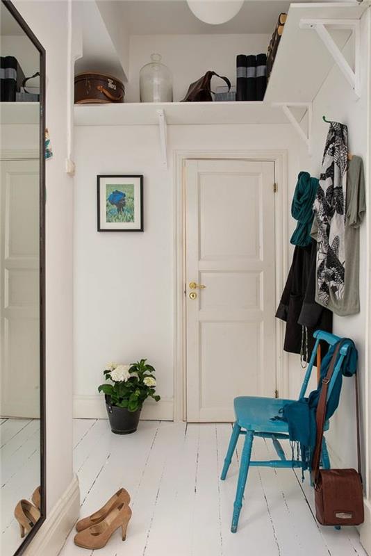 prieškambario apdaila su dideliu stačiakampiu veidrodžiu su juodu rėmu, medinė kėdė, nudažyta turkio spalva, grindys padengtos baltai dažyta mediena, juodas puodas su žydinčiu augalu baltomis gėlėmis