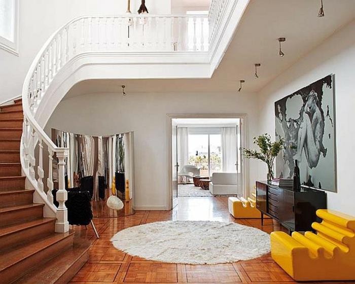 apvalus kilimas, rudos grindys, vingiuoti laiptai, geltoni foteliai, juoda komoda, milžiniška meninė tapyba