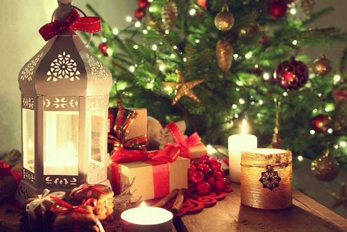 Kalėdiniai tapetai, medinis stalas, papuoštas baltu žibintu, romantiškos žvakės, dovanų dėžutės, papuoštos raudonomis juostelėmis, sausainiai ir papuošta žalia Kalėdų eglutė