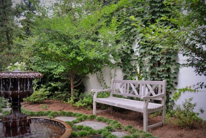 paprasta sodo dekoro idėja, fontanas, medinis suoliukas, akmeninis takas, vijokliniai augalai ant sienos, medžiai ir krūmai
