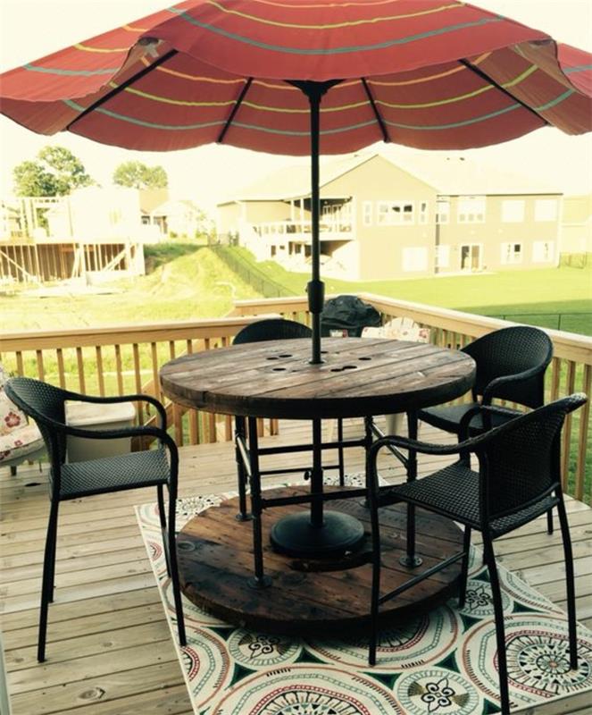 tik teras aranjmanı, eski ahşap masa makarası, ortasından geçen güneş şemsiyesi, siyah sandalyeler, oryantal desenli kilimler