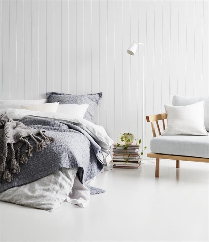 İskandinav esintili dekor, beyaz panelli duvar, beyaz ve gri nevresimler, gri zemin, beyaz minderli hafif ahşap kanepe, kitap yığını