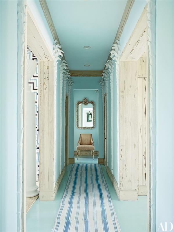 pastelinis žalias ir baltas koridorius, baltos ir mėlynos spalvos dryžuotas kilimas, smėlio spalvos fotelis fone ir baroko veidrodis