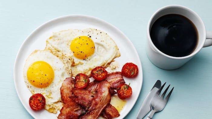 Skrudinti vyšniniai pomidorai, kiaušiniai ir šoninė baltoje lėkštėje su juoda kava, svorio netekimo meniu idėja, ketogeniniai pusryčiai