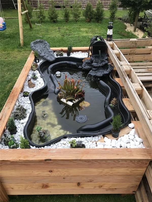 Çakıl taşları ve küçük yeşil bitkiler ve küçük musluk ile çevrili önceden oluşturulmuş siyah havza ile yükseltilmiş ahşap bahçe konteyner fikri