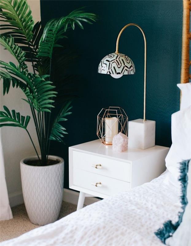Ideja za dekoriranje spalnice v skandinavskem dizajnu s steno v ozadju pavove modre barve, nočna omarica z belimi predali, belo posteljnino, sivo preprogo, svečko, zeleno palmo