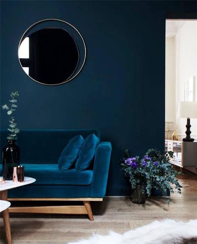 benzininės mėlynos sofos, atsiremiančios į tamsiai mėlyną sieną, idėja su kabančiu apvaliu veidrodžiu, šviesiu parketu, mediniu kavos staliuku, kilimėliu iš odos