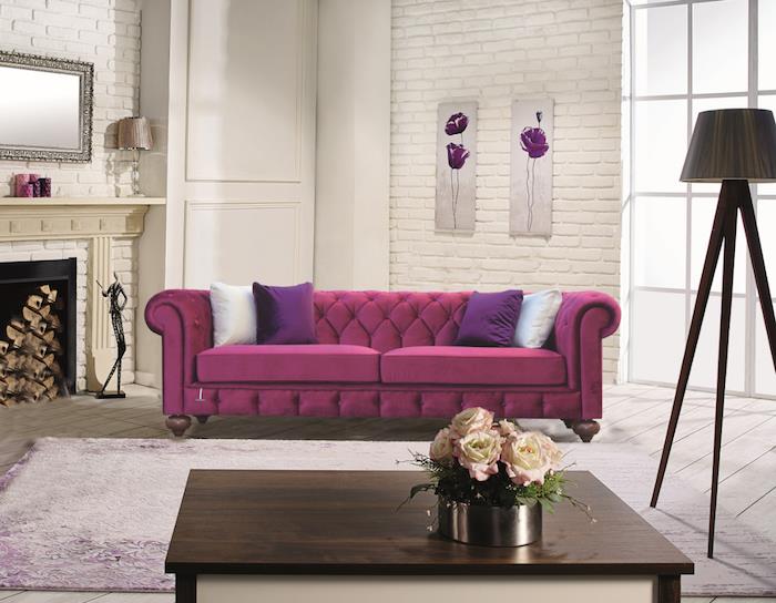 aviečių spalvos sofa baltoje svetainėje, plytomis dengtos sienos, baltas židinys, baltas ir purpurinis kilimas, rudas medinis kavos staliukas