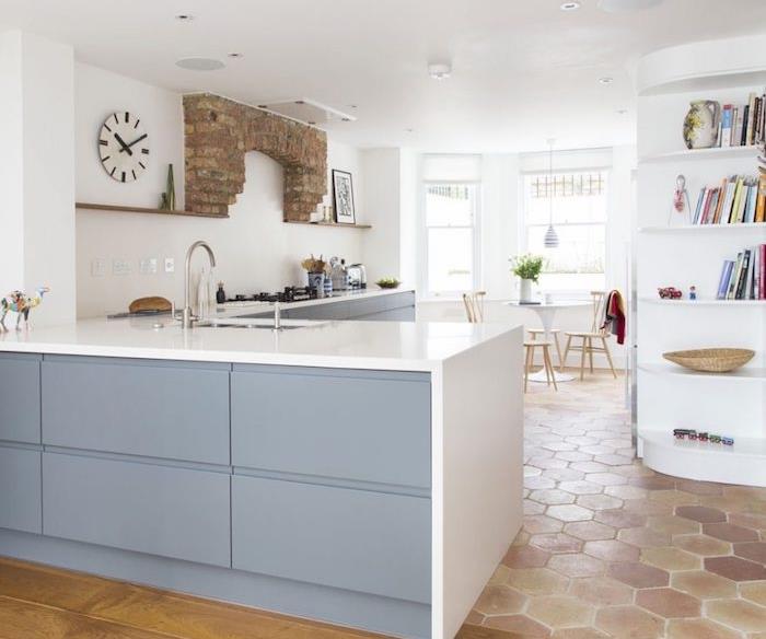 L formos virtuvės išdėstymas su pilkomis virtuvės spintelėmis ir baltu stalviršiu, maža balta knygų spinta, terakotos plytelių grindys, plytų akcentas