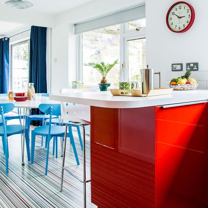 Yemek odasına açık bir mutfakta mavi ve kırmızı dekorasyon, beyaz tezgahlı kırmızı merkezi ada, beyaz masa ve mavi sandalyeler, beyaz arka plan