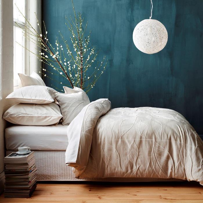 benzininis mėlynas miegamasis tamsių spalvų sienų dažais, baltas rutulinis pakabukas, balta patalynė, šviesus parketas, šūsnis žurnalų, dekoratyvinis medis