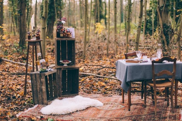 kaimiškos vestuvės miško aplinkoje, rytietiškas kilimas, medinis stalas ir kėdės, medinė dėžė, buteliukai ir gėlių buteliai