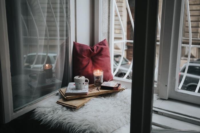 Kalėdinis fonas, higge tipo, sėdynės pagalvėlė prie lango krašto, medinis padėklas su knyga, puodelis karšto šokolado ir romantiškos žvakės, raudona pagalvėlė