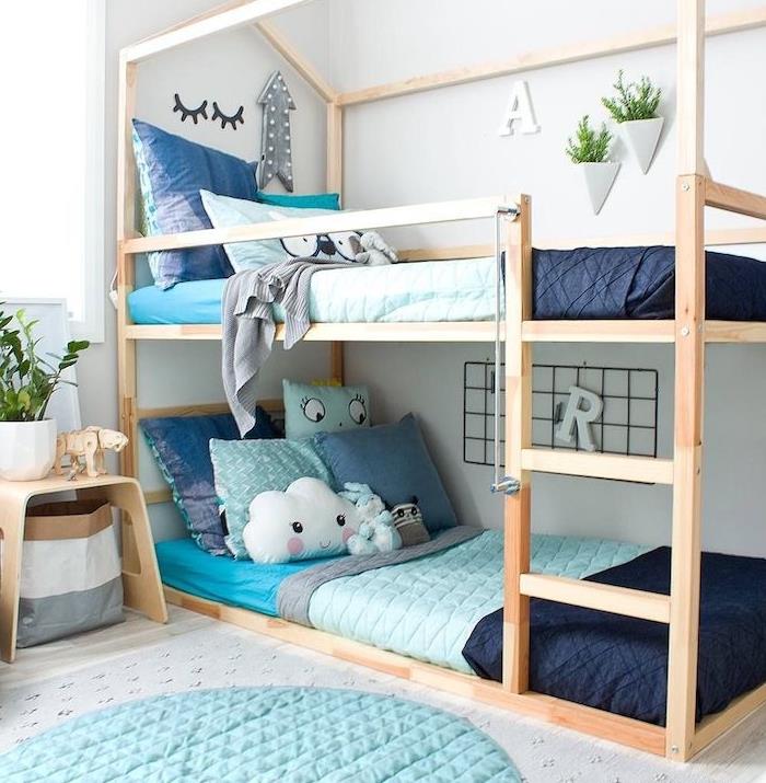 Skandinaviško stiliaus medinės dviaukštės lovos, taupančios vietą Montessori kambaryje, mėlyna patalynė, medinis naktinis stalas, mėlynas apvalus kilimas