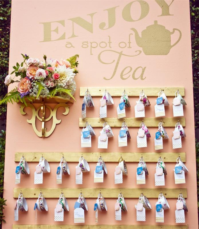 pavyzdinis vestuvių stalo planas, rožinis lašišos fonas, pakabinami arbatos puodeliai su gėlių puokšte šalia, arbatos pertrauka, mažos spalvotos etiketės