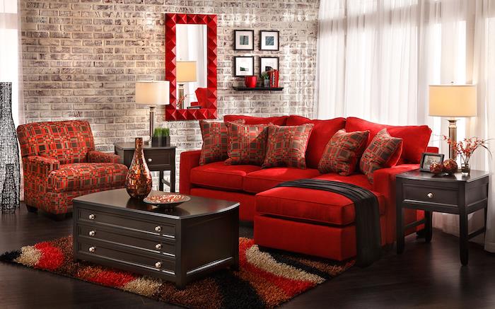 raudona kampinė sofa, raudonos ir rudos spalvos fotelis ir dekoratyvinės pagalvėlės, spalvingas kilimas, tamsiai rudas stalas ir parketas, plytų akcento siena