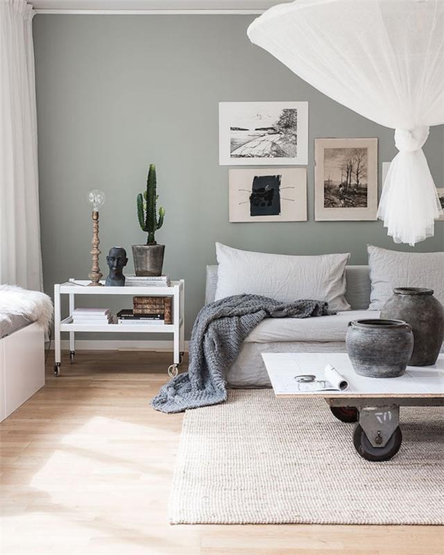 Nordijska dnevna soba s sivobelim kavčem, mizico na kolesih, v industrijskem slogu, svetlo sivo preprogo, sivo odejo, originalno belo visečo svetilko in stensko dekoracijo grafičnih modelov na sivo-zeleni steni celadon