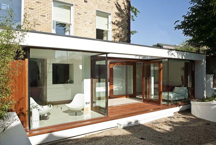 minimalistinio dizaino idėja - stiklo pailginimas su lova ir svetainės zona baltuose foteliuose, namo pratęsimas plokščiu stogu