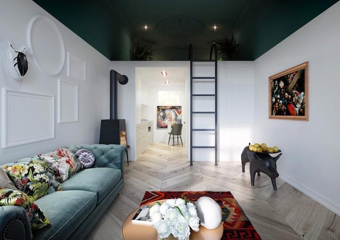 tamsiai žalios lubos ir baltos sienos, maža balta virtuvė, didelė povo mėlyna sofa, papuošta gėlėtomis pagalvėmis, spalvingas kilimas, sienos dekoruotos meno paveikslais