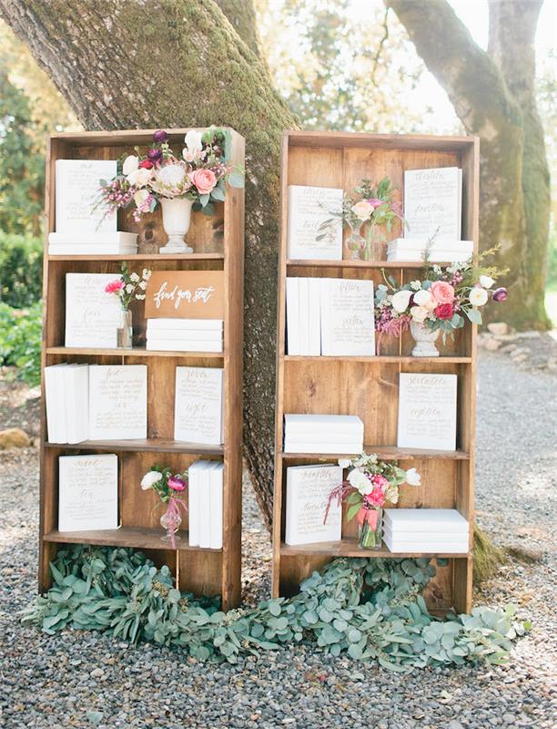 vestuvių stalo planas senose medinėse knygų spintelėse su baltomis knygomis, ant kurių viršelio užrašyti svečių vardai, gėlių puokštės baltose vazose