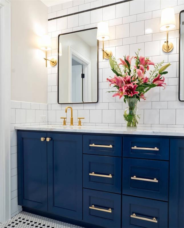 temno modra kopalniška omara v beli in modri kopalnici z belimi ploščicami, šopkom rož in medeninastimi poudarki