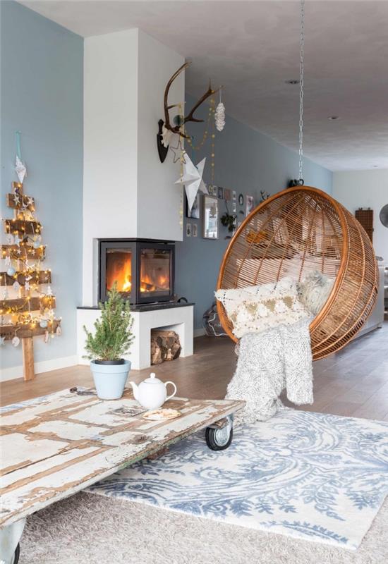 „Cocooning“ skandinaviškas gyvenamojo kambario baltos ir mėlynos pilkos spalvos dekoras, kuris kviečia atsipalaiduoti dėl savo kabančios rotango kėdės ir kalėdinės dekoracijos bei medinės eglutės, papuoštos mažomis šviesiomis girliandomis