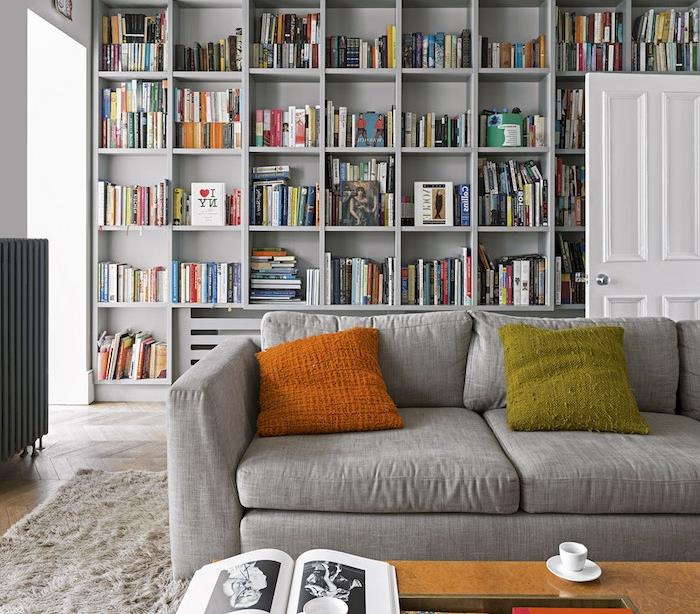 gri kitaplık saçak turuncu ve yeşil yastık, inci gri kanepe, yumuşak gri halı, ahşap sehpa ile modern oturma odası dekor fikri
