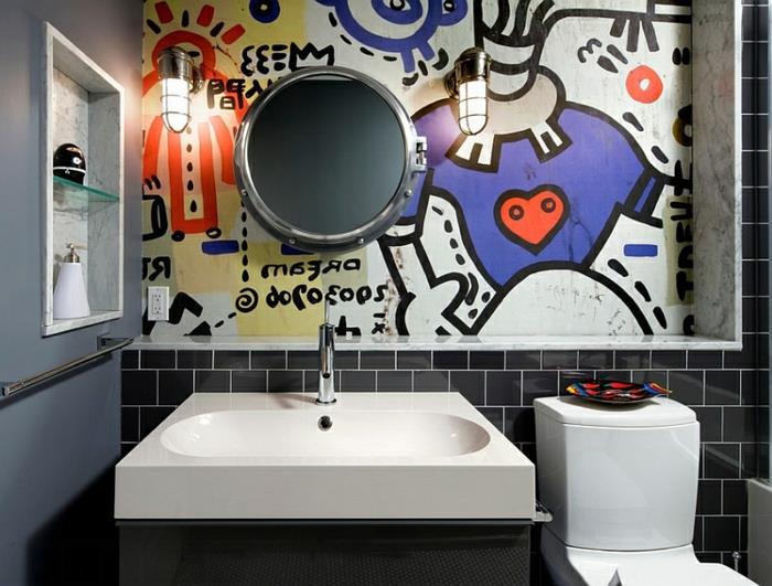 juodos metro tualeto plytelės, originalus sienų piešinys su spalvingu dizainu, apvalus veidrodis, baltas stalviršio praustuvas, pilka siena, du pramoniniai sieniniai žibintai