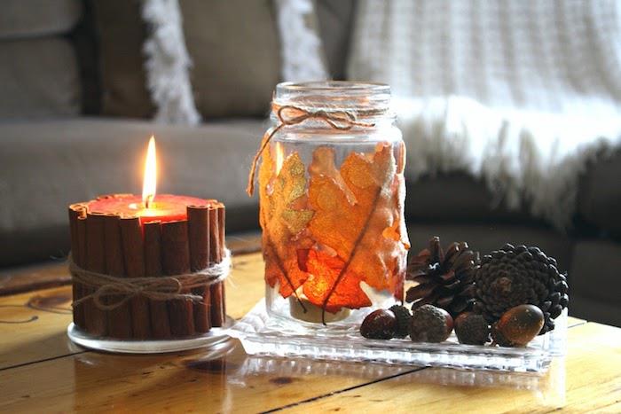 sveča, obdana s cimetovimi palčkami poleg prilagojenega steklenega kozarca z odpadlim listjem, borovimi storži in želodom v stekleni plošči