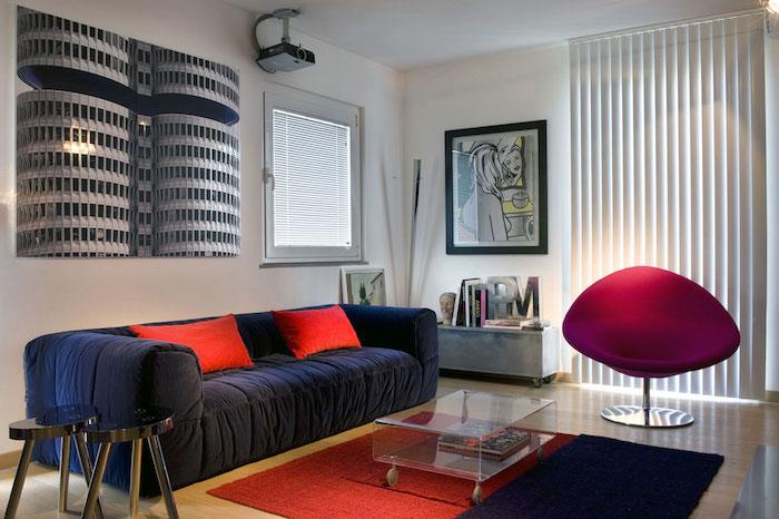 baltos sienos idėja, tamsiai mėlyna sofa, papuošta raudonomis pagalvėlėmis, aviečių spalvos fotelis, plastikinis stalas, mėlynas ir raudonas kilimas, originali sienų apdaila