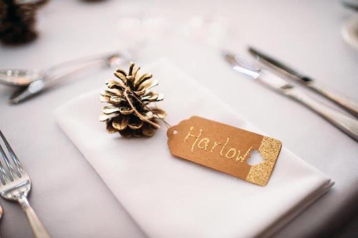 poceni poročni dekor, borov storž prebarvan v zlato barvo z zlato etiketo iz kraft papirja na belem prtičku