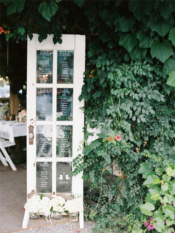 orijinal bir nikah masası planı için fikir, cam karolarda misafirlerin isimlerinin yazılı olduğu eski bir kapı, beyaz keçe camla yazılmış liste, doğal çerçeve